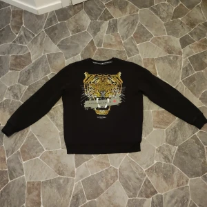 Svart tröja med tigertryck Alessandro Zavetti - Svart sweatshirt från Alessandro Zavetti med ett stort, detaljerat tigertryck framtill. Tröjan har rund hals, långa ärmar och ribbade muddar. Snygg broderad design med färger i gult, orange och vitt samt en grå detalj med logga.