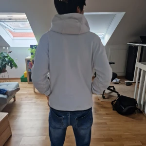 Vit Nike vindjacka med huva - Säljer en vit Nike vindjacka med svart dragkedja och svarta detaljer. Jackan har huva, lång ärm och en snygg V-formad design framtill. Perfekt för dig som gillar sportig stil och vill ha något lätt och bekvämt.