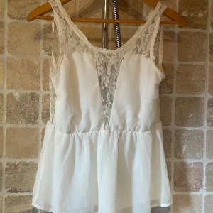 Vit spetslinnetopp från Vero Moda - Supersöt vit linne-topp från Vero Moda med spetsdetaljer upptill och längs axelbanden. Toppen har en v-ringad spetspanel framtill och ett flowy, lätt material nedtill. Perfekt för sommarens alla tillfällen och ger en romantisk vibe.