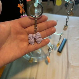 Lila rosett nyckelring - Supergullig nyckelring med en lila rosett i mjukt material. Rosetten hänger i en silvrig kedja och ring, perfekt att fästa på väskan eller nycklarna för en söt detalj. Enkel men ändå unik accessoar som lyfter din stil.