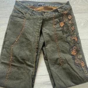 Broderade jeansbyxor Night Queen - Unika mörkgröna jeansbyxor från Night Queen med snyggt broderi och paljetter i brons på ena benet. Byxorna har raka ben, normal passform och coola dekorativa sömmar. Perfekta för dig som vill sticka ut med detaljer och mönster.