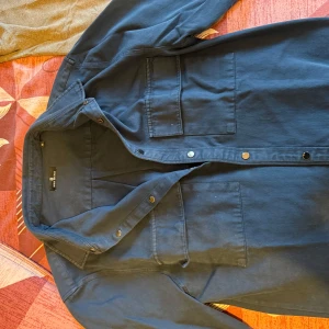 Mörkblå overshirt från Boris Becker XL - Snygg mörkblå overshirt från Boris Becker i storlek XL. Skjortan har två stora bröstfickor med lock, klassisk krage och tryckknappar framtill. Perfekt att bära öppen över en t-shirt eller stängd som en lätt jacka. Materialet känns slitstarkt och är i bomull.