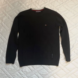 Svart stickad tröja Tommy Hilfiger - Svart stickad tröja från Tommy Hilfiger med logga på bröstet. Tröjan har rund halsringning, ribbade muddar och ett subtilt diamantmönster i stickningen. Perfekt för dig som gillar klassisk och stilren look.