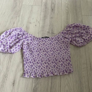Ljuslila blommig topp från Gina Tricot - Supersöt ljuslila topp från Gina Tricot med små lila blommor, puffiga korta ärmar och smockad kropp. Toppen har volangkant nertill och en fyrkantig urringning. Perfekt för dig som gillar romantisk och trendig stil.