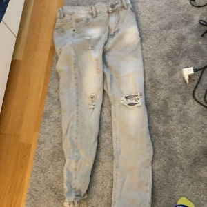 Ljusblå slitna jeans med hål - Säljer ett par ljusblå jeans med snygga slitningar och hål på ena benet. Jeansen har klassisk femficksdesign, normal passform och raka ben. Perfekta för en avslappnad och trendig look.