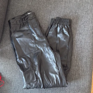 Svarta cargopants i skinnimitation FB Sister - Snygga svarta cargopants från FB Sister i skinnimitation. Byxorna har hög midja, resår vid benslut, stora fickor på sidorna och dragkedja i gylfen. Perfekta för en edgy streetstyle-look.