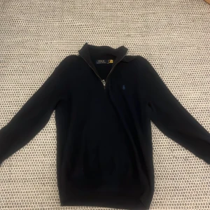 Mörkblå stickad half zip från Polo Ralph Lauren - Snygg mörkblå stickad tröja från Polo Ralph Lauren med halv dragkedja och klassisk broderad logga på bröstet. Tröjan har lång ärm och en stilren krage, perfekt för lager-på-lager-look. Passar dig som gillar tidlös och clean stil.