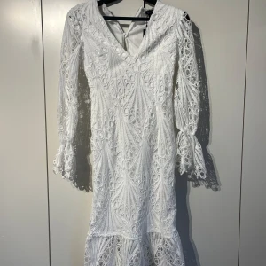 Vit spetsklänning med volangärm - Superfin vit klänning med v-ringning, långärmade spetsärmar och volangdetaljer vid ärmslutet. Klänningen har ett snyggt spetsmönster över hela och en figurnära passform. Perfekt för sommarens alla tillfällen! Dragkedjan är sönder men de går att fixa! 