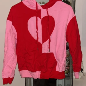Rosa och röd hoodie med hjärta - Säljer en färgstark hoodie i rosa och rött med ett stort hjärtmönster framtill. Tröjan har huva med snörning, magficka och långa ärmar. Perfekt statement-plagg för dig som gillar att sticka ut och sprida kärlek.