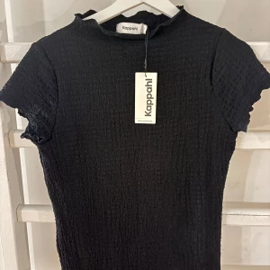 Svart ribbad topp från Kappahl - Superfin svart topp från Kappahl med smockad struktur och lätt volang vid ärmslut och halsringning. Kortärmad och i stretchigt material.