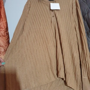 Beige ribbad långklänning med krage - Säljer en beige långklänning med ribbat mönster och snygg krage. Klänningen har långa, vida ärmar och knappar framtill. Materialet är mjukt och flowy, perfekt för dig som gillar en chill och avslappnad stil.