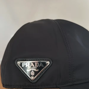 Svart keps från Prada - Snygg svart keps från Prada med klassisk böjd skärm och justerbar rem baktill. Kepsen har Pradas ikoniska metallogo framtill och är tillverkad i glansig nylon som ger en lyxig känsla. Perfekt accessoar för att lyfta din outfit. Kepsen är helt ny och inte använd