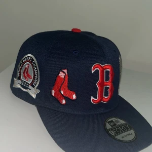 Boston Red Sox keps New Era 9FORTY - Mörkblå Boston Red Sox keps från New Era 9FORTY med broderade detaljer som röd B-logga, strumpor, World Series Champions-märke och guldtrofé. Justerbar snapback i rött och broderad text 'BOSTON' bak. Perfekt för basebollfans.