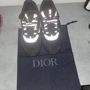 Svarta sneakers med reflex detaljer  - Snygga svarta sneakers med reflekterande detaljer. Skorna har snörning och en modern design med en kombination av syntetmaterial och mesh för extra komfort.