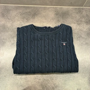 Mörkblå kabelstickad tröja från GANT - Kabelstickad mörkblå tröja från GANT med rund halsringning och broderad logga på bröstet. Tröjan är i mjuk bomull och har långa ärmar samt ribbade muddar. Perfekt för dig som gillar klassisk och stilren look.