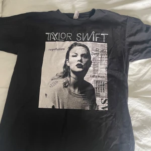 Taylor Swift svart t-shirt  - Svart Taylor Swift t-shirt från Fruit of the Loom med stort tryck på framsidan från Reputation-eran. Klassisk passform, rund hals och korta ärmar. Materialet är mjuk bomull och trycket har tidningsinspirerad design i svartvitt. Perfekt för Swifties!
