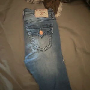 True Religion Joey Mid Rise Flare Jeans - Snygga blå jeans från True Religion, modell Joey Mid Rise Flare. Jeansen har medelhög midja, utsvängda ben och klassiska detaljer med kontrastsömmar och logga på bakfickan. Mjukt denimtyg med slitningar och kopparfärgade knappar. Byxorna har ej används. 