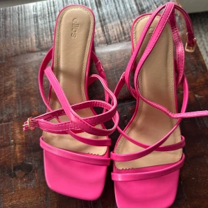 Rosa oanvända klackar från Ellos - Snygga sandaler i starkt rosa med fyrkantig tå och blockklack. Remmarna är tunna och går i kors över foten, med justerbart spänne vid ankeln. Aldrig använda! 