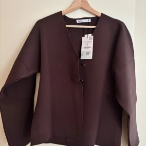 Brun cardigan jacka från Zara - Mörkbrun cardigan jacka från Zara i storlek L. Jackan har en stilren v-ringning, två guldiga knappar framtill och är tillverkad i ett mjukt, stickat material. Perfekt för dig som gillar minimalistisk och clean stil.