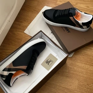 Burberry sneakers med klassiskt rutmönster - Snygga svarta sneakers från Burberry med den ikoniska rutan i beige, svart och vitt på hälen. Skorna har vita platta sulor, svarta snören och detaljer i syntet och skinn. Perfekta för dig som vill ha en stilren men ändå lyxig look.//// passar 43
