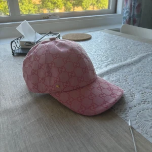 Rosa Gucci keps - Säljer en rosa keps från Gucci med ikoniskt GG-mönster över hela kepsen. Justerbar rem baktill i rosa läder och klassisk böjd skärm. Kepsen är tillverkad i bomull och har en lyxig känsla som sticker ut i mängden.