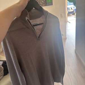 Grå merinoulltröja från These Glory Days - Säljer en stilren grå långärmad tröja från These Glory Days i mjuk merinoull. Tröjan har half zip-dragkedja vid halsen och en klassisk passform som funkar till många outfits. Perfekt för dig som gillar en clean och tidlös look.