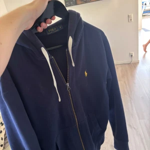 Marinblå hoodie från Polo Ralph Lauren - Snygg marinblå hoodie från Polo Ralph Lauren med dragkedja, huva och vita snören. Klassisk gul logga broderad på bröstet och stora fickor framtill. Perfekt för dig som gillar stilren streetwear med premiumkänsla.