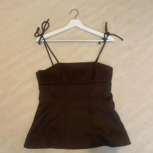 Brun linnetopp med knytband - Supersnygg brun linnetopp med smala axelband som knyts på axlarna. Toppen har en rak, något figurnära passform och är lätt utsvängd nertill. Perfekt att styla med jeans eller kjol för en trendig look.