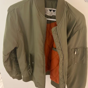 Olivgrön bomberjacka från Junkyard - Snygg olivgrön bomberjacka från Junkyard med orange quiltat foder. Jackan har dragkedja framtill, fickor med tryckknapp och en ärmficka med dragkedja. Klassisk ribbad mudd vid ärmslut och midja. Perfekt för dig som gillar streetstyle.