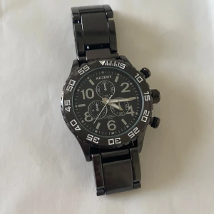 Svart klocka från Akzent med länkarmband - Stilren svart klocka från Akzent med robust länkarmband i metall. Urtavlan är svart med vita siffror och detaljer, samt tre mindre urtavlor för extra funktioner. Klockan har tydliga knappar på sidan och sportig design. Helt ny med nya batteri 