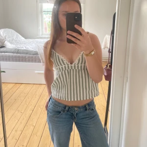 Randig topp med spetsdetaljer - Säljer en söt randig linnetopp med gröna och vita vertikala ränder. Toppen har smala axelband, v-ringning och fina spetsdetaljer längs urringningen. Croppad modell som ger en avslappnad vibe, perfekt för varma dagar. Den är köpt från en som syr egna toppar