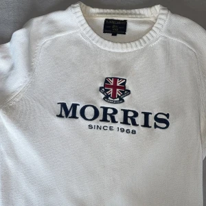 Vit stickad tröja från Morris Gents - Vit stickad tröja från Morris Gents med broderad logga och brittisk flagga på bröstet. Klassisk rund halsringning och ribbade muddar. Perfekt för dig som gillar stilren och preppy look.