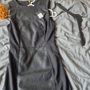 Svart kort klänning från New Look - Snygg svart kort klänning från New Look i storlek 34. Klänningen har paneler i skinnimitation och en guldig dragkedja bak. Ärmlös och figurnära modell som ger en cool och stilren look. Perfekt för dig som gillar edgy vibes.