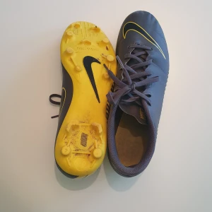 Nike fotbollsskor i grått och gult - Snygga fotbollsskor från Nike i mörkgrått med gula detaljer och sula. Skorna har snörning och en klassisk Nike-logga i gult på ovansidan. Ovandelen är i syntetmaterial och sulan har dobbar för bra grepp på planen.