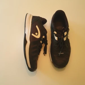 Svarta inomhussskor från Head - Svarta tennisskor från Head med vita detaljer och snörning. Ovandelen är i mesh och syntet för bra andningsförmåga och komfort. Sulan har fiskbensmönster för extra grepp på banan. Perfekta för dig som vill ha stabilitet och stil på tennisplanen.