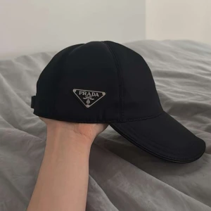 Svart keps från Prada i nylon - Snygg svart keps från Prada med klassisk böjd skärm och ikonisk metallogo på sidan. Kepsen är tillverkad i slät nylon och har justerbar rem baktill för perfekt passform. En stilren accessoar som lyfter vilken outfit som helst. Priset är ej hugen i sten, tveka inte med att ställa frågor!😄