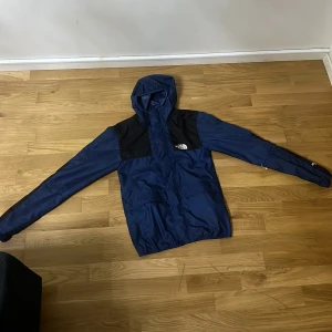 Blå och svart vindjacka The North Face - Snygg blå och svart vindjacka från The North Face med huva och dragkedja framtill. Jackan har logga på bröstet, svarta partier över axlar och ärmar samt praktiska fickor. Perfekt för blåsiga dagar och outdoor vibes. Det finns en fläck på baksidan av jackan men det går säkert bort i tvätten. Skriv gärna vid intresse!