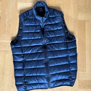 Blå dunväst från Uniqlo (Ultra Light Down) - Säljer en marinblå dunväst från Uniqlo och det är modellen Ultra Light Down. Västen är quiltad med horisontella sömmar, har hög krage och dragkedja framtill. Den är superlätt och perfekt för lager-på-lager. Materialet är glansigt och vattenavvisande.      Hör av dig ifall du har minsta lilla fråga om västen! 