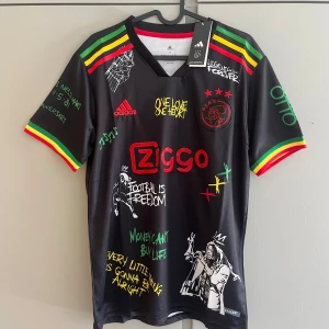 Ajax x Bob Marley Adidas fotbollströja - Unik svart Ajax fotbollströja från Adidas med Bob Marley-tema. Färgglada detaljer i rött, gult och grönt på ärmarna, tryck med Ziggo-logga, Ajax-märke och Bob Marley-motiv på ryggen. Coola citat och graffiti-liknande tryck över hela tröjan. Tillverkad i lätt och ventilerande material.