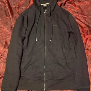 Svart hoodie från BOSS med dragkedja - Svart hoodie från BOSS med diskret logga på bröstet. Tröjan har hel dragkedja, huva med snörning och två fickor framtill. Tillverkad i mjuk bomull och har långa ärmar. Perfekt för en chill och stilren look.