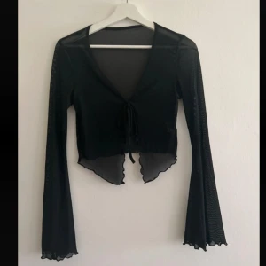 Svart mesh bolerotopp med knytband - Säljer en svart transparent bolerotopp i mesh med långa trumpetärmar och vågig kant nertill. Toppen har v-ringning och knytband framtill för en snygg detalj. Perfekt att styla över linne eller klänning för en trendig look.