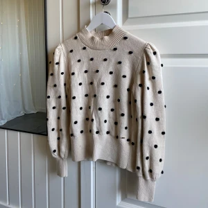Beige stickad prickig tröja - Superfin beige stickad tröja med svarta prickar och puffiga långärmar. Hög ribbad halsringning och breda muddar ger en trendig look. Perfekt för dig som gillar lekfulla detaljer. 