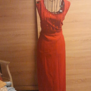 Röd långklänning XLNT by KappAhl - Snygg röd långklänning från XLNT by KappAhl i storlek 54. Klänningen har ärmlös design, rund halsringning och dekorativa paljettdetaljer framtill. Perfekt för dig som vill sticka ut med en klassisk och elegant look.