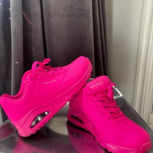 Chockrosa sneakers från Skechers - Färgstarka sneakers från Skechers i en riktigt chockrosa nyans. Skorna har en chunky sula med synlig luftbubbla, snörning och rund tå. Perfekta för dig som vill sticka ut och addera färg till din outfit.