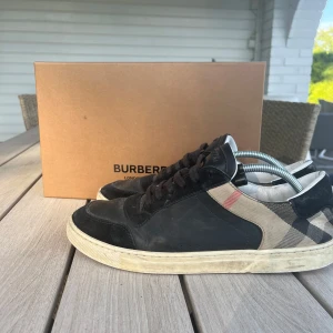 Burberry sneakers med klassiskt rutmönster - Snygga sneakers från Burberry med svart skinn och mocka framtill samt den ikoniska beigea och svarta rutan på sidorna. Skorna har svarta snören, vit sula och diskret Burberry-logga på plösen. Perfekta för dig som vill ha en stilren och lyxig look. Köpt från cettire för 5000 har kvitto och bevis ändast skor förkommer, om inte ext pengar läggs in 