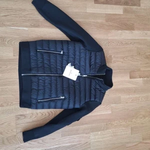 Svart Moncler cardigan herr - Snygg svart pufferjacka från Moncler med quiltad front och släta ärmar. Jackan har två dragkedjefickor, ribbade muddar och klassisk Moncler-logga på ärmen. Perfekt för dig som vill ha en stilren och exklusiv look i höst och vinter.