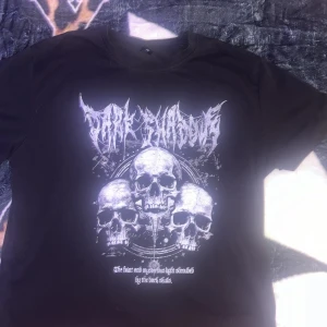 Svart band-tshirt med dödskallar - Svart t-shirt med tryck av tre dödskallar och bandlogga i vitt framtill. Klassisk rund hals och korta ärmar. Perfekt för dig som gillar metal eller rock och vill ha en edgy stil.
