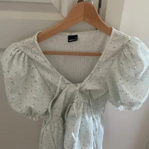 Blommig blus med puffärm Gina Tricot - Supersöt ljus blus från Gina Tricot med små blommor, puffiga korta ärmar och knyt framtill. V-ringad och lätt rynkad i midjan för en feminin siluett. Perfekt för vår och sommar när du vill ha en romantisk vibe.
