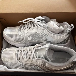 New Balance 530 vita sneakers - Snygga New Balance 530 sneakers i vitt med metallic-detaljer. Ovandel i mesh och syntet för en sportig vibe, med klassisk N-logga på sidan. Skorna har snörning och en chunky sula med svart och blått mönster undertill. Perfekta för en clean och modern look.