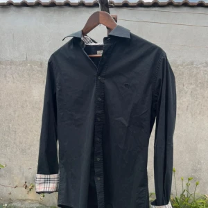 Svart skjorta från Burbery med rutiga detaljer - Stilren svart skjorta från Burbery med klassiska rutiga detaljer på insidan av manschetter och krage. Skjortan har knappar framtill, lång ärm och diskret broderad logga på bröstet. Tillverkad i bomull med lite elastan för extra komfort. Köp inte direkt skriv till mig först!!!!
